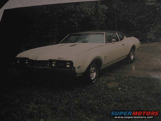 White coupe with a black vinyl top, parked on grass. Visible grille, headlights, and side emblem