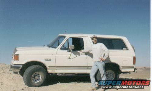 Man in white sweatshirt and jeans stands by a white 1990 Ford Bronco in a desert landscape