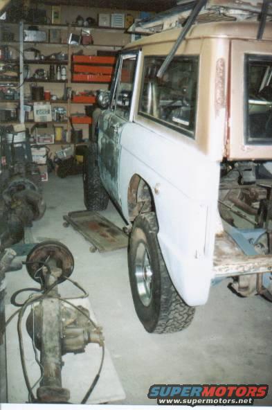 Ford Bronco body in garage, primer on front fenders, tan hardtop. Parts and tools on shelves in background