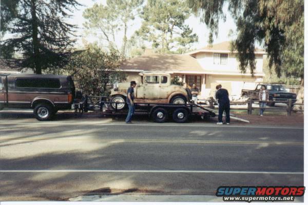 Rusty 1930s coupe on a trailer, towed by a brown pickup truck