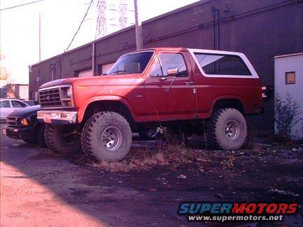 Red Ford Bronco with large tires parked next to a building, front-left view