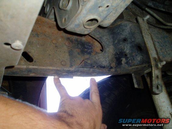Close-up of rusty vehicle frame and suspension parts, with fingers pointing to a section