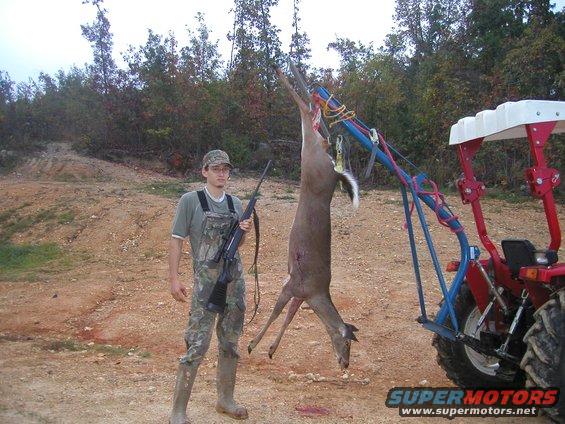 Hunter with rifle stands next to a deer hanging from a tractor lift