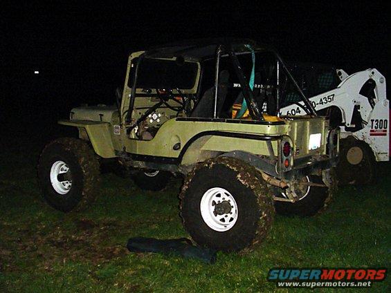 Olive green Jeep CJ-5 with a roll cage, mud-caked tires, and a white skid steer loader behind it