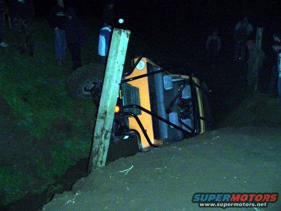 Orange off-road vehicle tipped on its side next to a wooden post, with people watching in the background at night