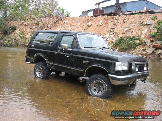 Black Ford Bronco driving through a shallow stream, tires submerged. Some mud on the body. Text on windshield reads 'FULL THROTTLE'