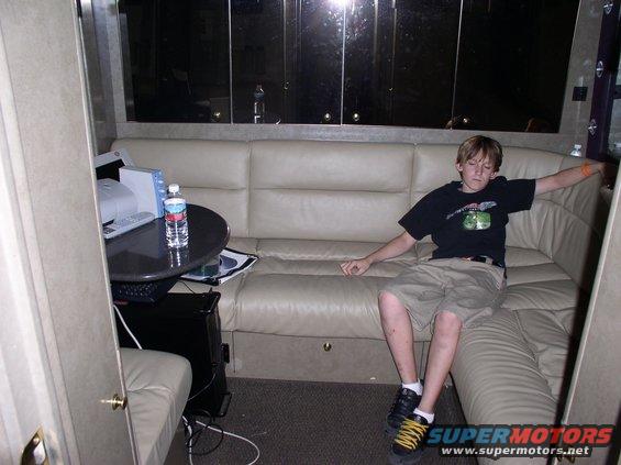 Boy sitting on a couch inside an RV, with a table and computer equipment nearby