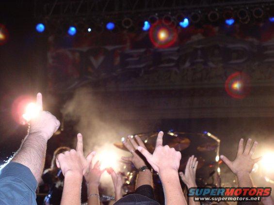 Concert crowd with hands raised in the air, stage lights and smoke visible