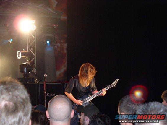 Guitarist with long hair playing an electric guitar on a stage with bright lights and Marshall amplifiers