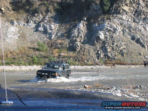 1992 Ford Bronco driving through shallow river, rocky hillside in background
