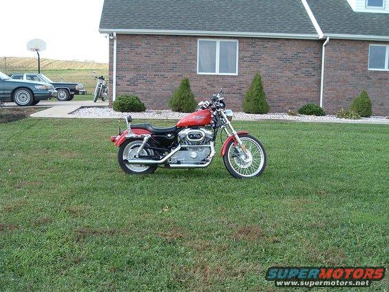 Red Harley-Davidson motorcycle parked on grass in front of a brick building; two cars and another motorcycle are visible in the background
