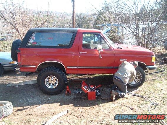 1987 Ford Bronco with a person working underneath it, spare tire on the back