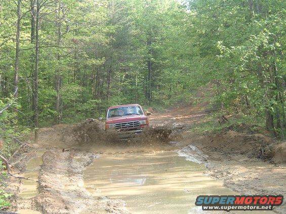 Red 1987 Ford Bronco splashing through mud on a wooded trail