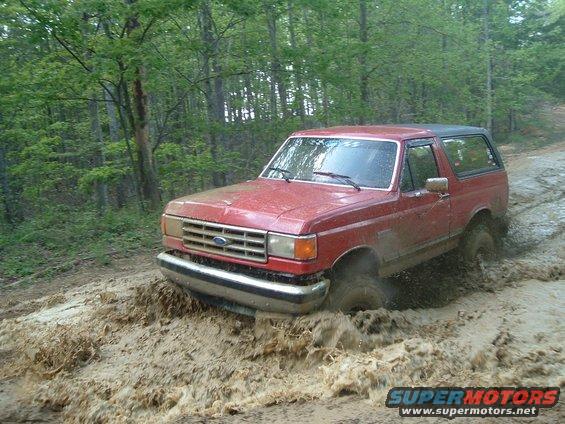 Red 1987 Ford Bronco driving through mud on a wooded trail