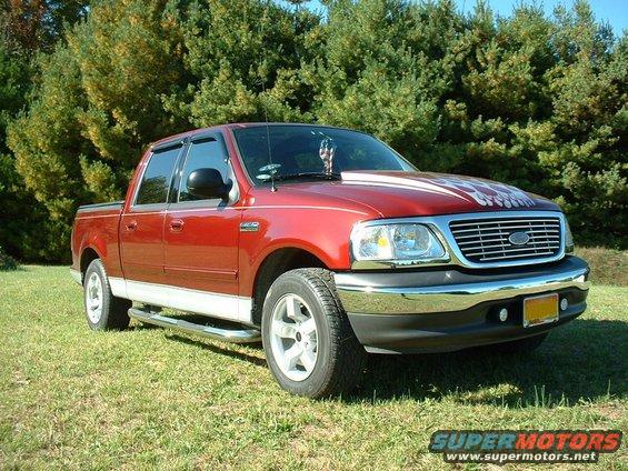 Red Ford F-150 Supercrew pickup truck parked on grass with trees in background. Has white racing stripes on hood