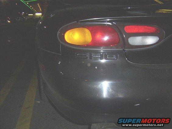 Dark sedan's rear, showing taillight and 'SHO' badging. The taillight has a yellow turn signal and red brake light