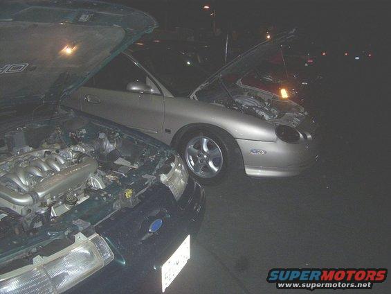 Two cars, one dark green and one silver, parked at night with hoods open. The silver car looks like a 1998 Mercury Sable