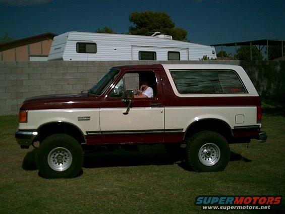 Two-tone red and cream Ford Bronco parked on grass with a trailer behind a wall