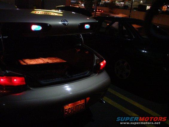 Rear view of a car with trunk open, showing a box inside. Another dark car is parked next to it