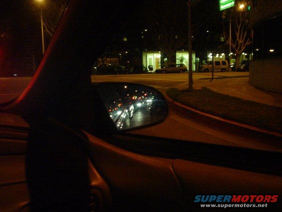 Night driving view from inside a car, showing the side mirror reflecting traffic lights and headlights