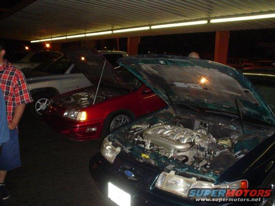 Several cars parked with hoods open, showing engines. One is dark green, another is red, and a silver truck is behind them