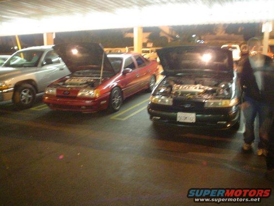 Two sedans, one red and one dark green, parked side-by-side with hoods open. A silver SUV is parked to the left