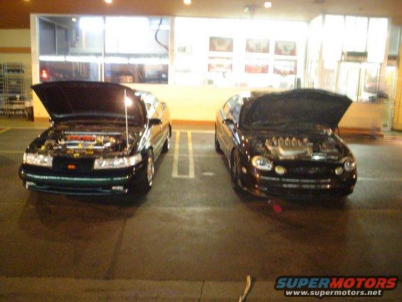 Two dark sedans parked side-by-side, both with hoods open showing their engines
