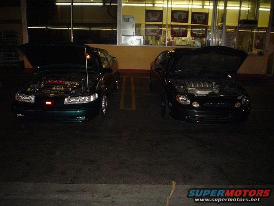 Two sedans parked at night, both with hoods open. One is dark green, the other is black