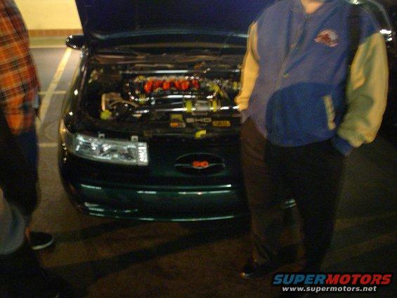 Front of a dark green Ford Taurus SHO with hood open, showing the engine. Two people stand nearby