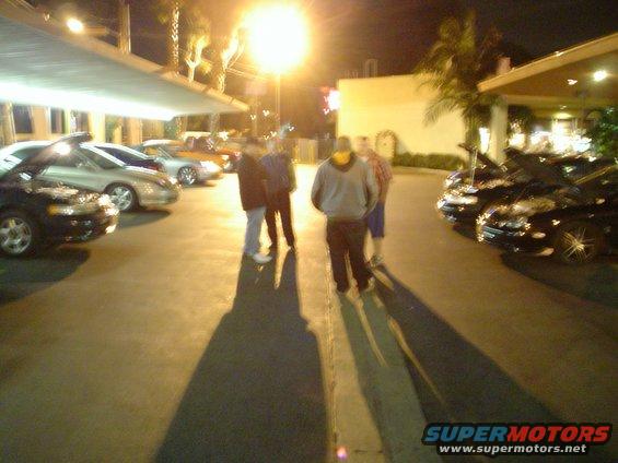 People standing in a nighttime parking lot with cars under bright lights