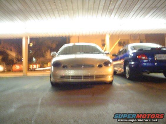Gold sedan parked under a covered area, front view. Another blue car is to the right