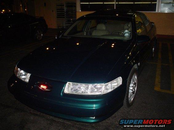 Dark green sedan with a red SHO badge on the grille. Another dark vehicle is parked to the left