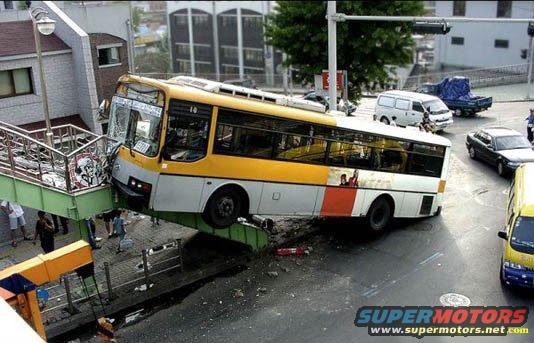 Yellow and white bus crashed onto a pedestrian overpass, front end damaged, side windows intact