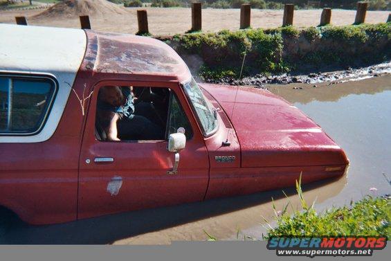 1978 Ford Bronco partially submerged in muddy water, driver visible inside