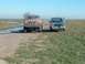 1987 Ford Bronco - Two trucks are parked on a dirt road with puddles and grassy areas under a clear blue sky.