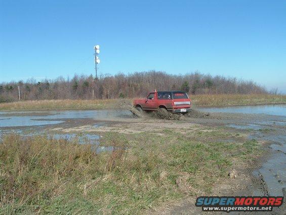 Red SUV kicking up mud while driving through a muddy field. Black roof, clear sky