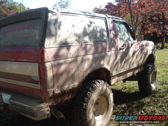 Red and white 1987 Ford Bronco covered in mud, lifted with large tires, viewed from the right rear