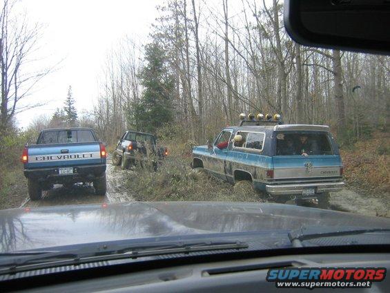 Blue Chevrolet K5 Blazer and a blue Chevrolet pickup truck driving on a muddy trail through trees