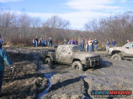 Two pickup trucks stuck deep in mud, with a group of people watching. The front truck is a light-colored pickup, heavily covered in mud