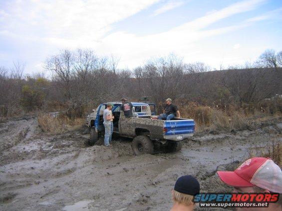 Mud-splattered blue pickup truck stuck in deep mud with people around it