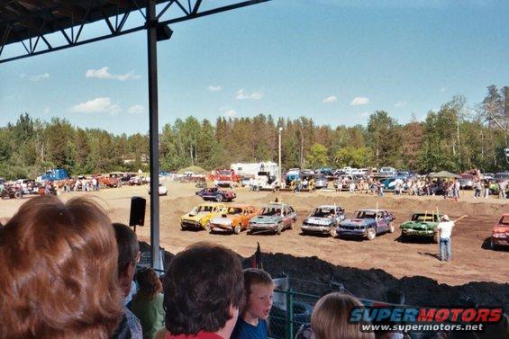 Demolition derby cars lined up on dirt track under a covered grandstand, with spectators in foreground