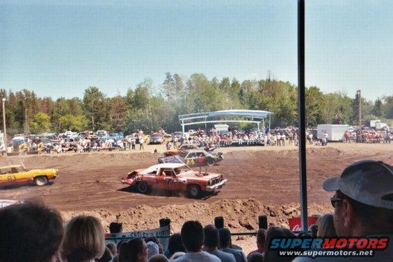 Demolition derby event with several cars on a dirt track, spectators in the background