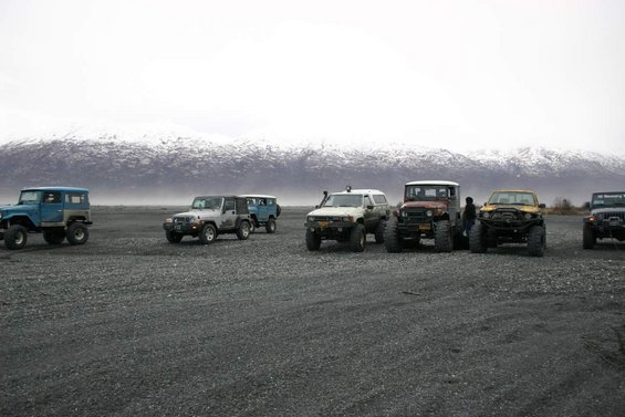 Several off-road vehicles including Jeeps and older SUVs parked on a gravel plain with snow-capped mountains in the background