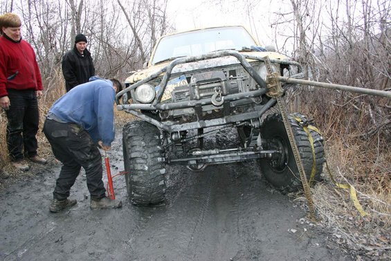 Yellow off-road vehicle stuck in mud, front bumper with winch, high-lift jack attached to tire