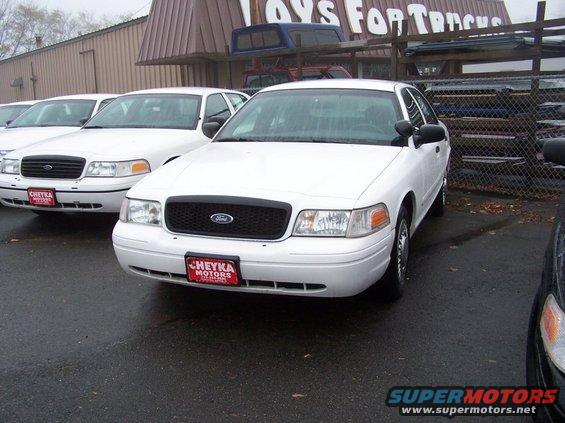 Front view of a white 2001 Ford Crown Victoria police car with a black grille and a 'CHEYKA MOTORS' license plate frame. Another similar car is parked to the left