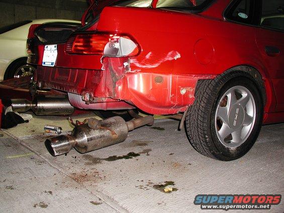 Red car's rear bumper is damaged, exhaust pipe detached. Another car is visible