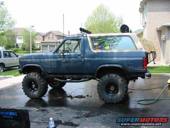 Blue Ford Bronco with large tires and roof-mounted lights, parked on a wet driveway