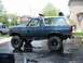 1986 Ford Bronco - A lifted blue Ford Bronco with off-road tires and accessories is parked on a wet surface, with a silver SUV in the background.