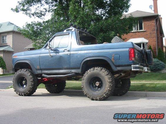 Blue Ford Bronco with large off-road tires, lifted suspension, and light bar on roof. Spare tire mounted on rear