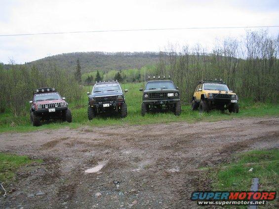 Four SUVs parked on a muddy dirt road with trees and a hill in the background